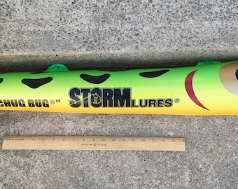May include: A large, green and yellow fishing lure with black spots and the text "CHUG BUG" and "STORM LURES". The lure has a large eye and a red mouth design. A wooden ruler is next to the lure.