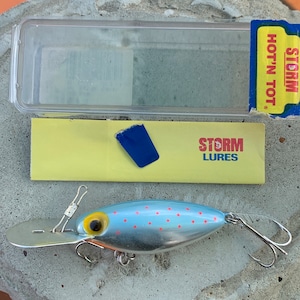 Könnte beinhalten: Ein Storm Hot 'N Tot Angelköder mit silbernem Körper, blauem Oberteil mit roten Punkten und gelbem Auge. Der Köder hat eine silberne Metalllippe und zwei Drillingshaken. Die Verpackung enthält eine transparente Kunststoffbox und eine gelbe Karte mit dem Text "STORM LURES".