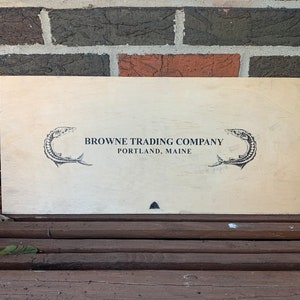 Puede incluir: Un letrero rectangular de madera con el texto "BROWNE TRADING COMPANY PORTLAND, MAINE" en negro. Dos peces estilizados flanquean el texto. El letrero tiene una veta de madera clara y natural.
