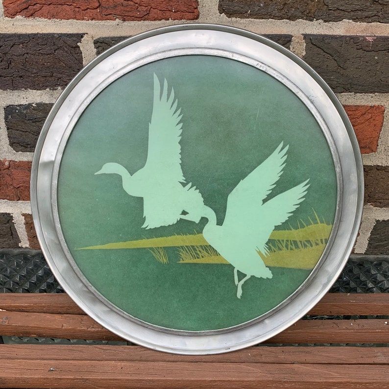 May include: Round metal tray with a green glass insert featuring a silhouette of two flying ducks. The ducks are light green, and the background has a green and yellow landscape design. The tray has a silver metal rim.