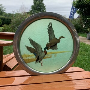 May include: Round metal tray with a painted scene of two ducks in flight over a marsh. The ducks have brown, green, and blue feathers. The background is a light green color. The tray has a silver-colored metal rim.