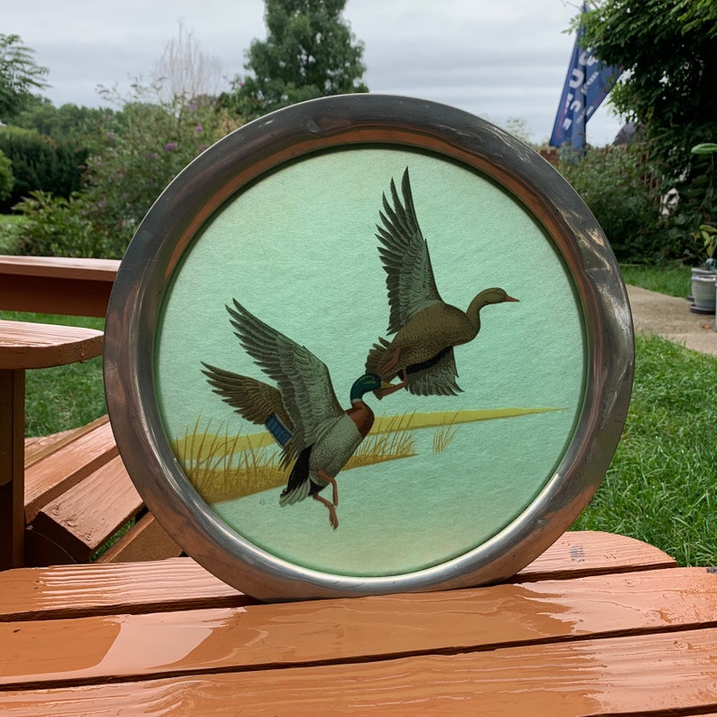 May include: Round metal tray with a painted scene of two ducks in flight over a marsh. The ducks have brown, green, and blue feathers. The background is a light green color. The tray has a silver-colored metal rim.