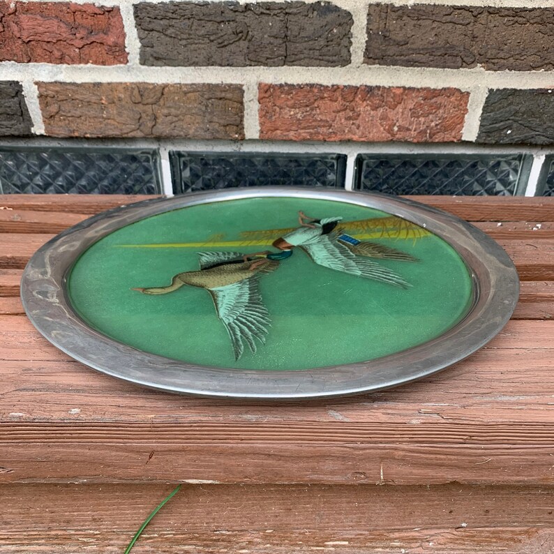May include: Oval serving tray with a silver-colored rim. The tray's interior features a green background with two ducks in flight. The ducks are depicted with detailed feathers and are painted in shades of brown, white, and blue.