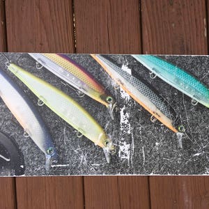 Könnte beinhalten: Eine Sammlung von Angelködern in verschiedenen Farben, darunter Silber, Gelb und Blau, angeordnet auf einer schwarz-weißen strukturierten Oberfläche. Jeder Köder hat einen Haken und ist zum Fangen von Fischen bestimmt.
