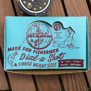 Könnte beinhalten: Eine Vintage-Angelkiste mit dem Text "MADE FOR FISHERMEN Dial-a-Shot 4 SINKER WEIGHT SIZES" in Rot und Weiß auf hellblauem Hintergrund. Die Box zeigt eine Grafik eines Fischers und eines Fisches.
