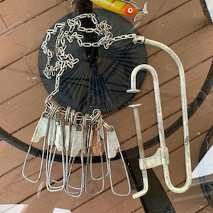 Puede incluir: Un surtido de aparejos de pesca antiguos, que incluye una cadena con múltiples clips metálicos, una abrazadera metálica y un señuelo de pesca colorido con una hélice. Los artículos están dispuestos sobre una superficie de vidrio.