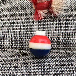Pode incluir: Uma rolha de garrafa decorativa redonda, vermelha, branca e azul, com uma parte superior branca. A rolha está sobre uma superfície texturizada, cinza escuro e bege. Um elemento decorativo vermelho e bege está no fundo.