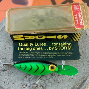 Könnte beinhalten: Ein grüner und schwarzer Angelköder mit gelbem Auge und silbernen Haken. Der Köder befindet sich in seiner Originalverpackung mit dem Text "Quality Lures... for taking the big ones... by STORM."