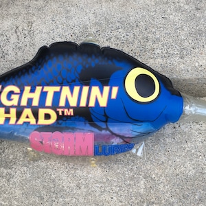 Könnte beinhalten: Ein blau-schwarzer Lightning Shad-Angelköder mit einer durchsichtigen Kunststofflippe. Der Köder hat ein gelbes Auge und den Text "LIGHTNIN' SHAD" in Gelb. Die Verpackung enthält den Text "STORM LURES" in Pink.