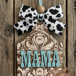 May include: A wooden tag-shaped ornament featuring a dark brown laser-cut damask pattern on a light wood background. The word MAMA is displayed in raised, light blue block letters. A cow-print ribbon bow and a wooden bead hang from a light blue cord.