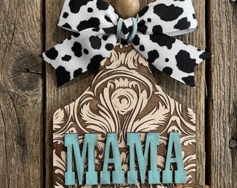 May include: A wooden tag-shaped ornament featuring a laser-cut damask pattern and the word MAMA in raised teal letters. The ornament is topped with a natural wooden bead and a white ribbon with a black cow print pattern, suspended by a light blue cord.