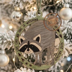 May include: A circular wooden ornament featuring a gray raccoon illustration in the center. The frame includes the text "LIVE LIKE NO ONE IS WATCHING" and "JIMOTHY 2026" with small paw print accents. It hangs from a green string.