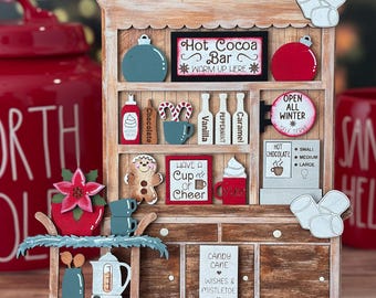 May include: A wooden hutch display featuring miniature holiday-themed items including a Hot Cocoa Bar sign, gingerbread man, candy canes, syrup bottles, and a small cart with a kettle, all decorated with red, white, and green accents.
