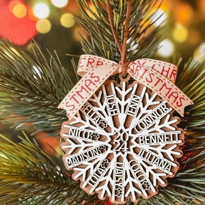Może przedstawiać: Drewniana ozdoba świąteczna w kształcie płatka śniegu z imionami i odciskami łap, zawieszona na choince. Ozdoba ma rok 2024 i słowo "Christmas" na wstążce. Imiona to Bella, Nico, Josh, Jenn, Connor, Bennett, Madison, Lola, Daisy, Coco i Juliana.