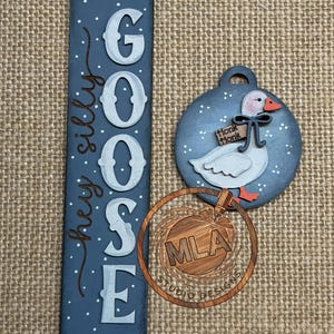 May include: Wooden goose-themed decorations. A blue vertical sign says "GOOSE" with "hey silly" in script. A round ornament features a goose with a "Honk Honk" sign. A wooden logo is also included.