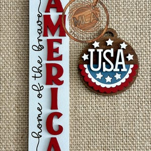 May include: A patriotic wooden sign with the words "AMERICA home of the brave" in red and black lettering. A wooden ornament with "USA" and stars is also present. The sign and ornament are on a burlap background.