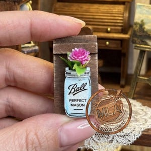 May include: A miniature wooden wall plaque featuring a light blue mason jar illustration with the text Ball PERFECT MASON printed in black. A small pink flower with green leaves is attached to the top of the jar, creating a decorative wall hanging.