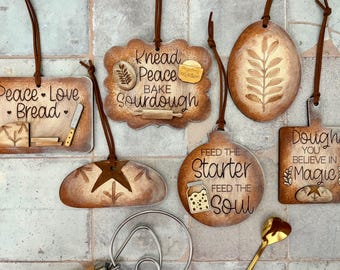 May include: A collection of wooden ornaments with a bread-making theme. Each ornament features a unique shape and design, including text like "Peace, Love, Bread" and "Feed the Starter, Feed the Soul." The ornaments are connected with brown cords.