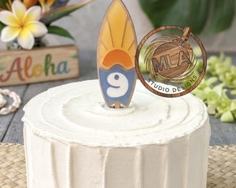 Puede incluir: Un pastel redondo con glaseado blanco presenta un adorno en forma de tabla de surf. El adorno muestra un gráfico de sol amarillo sobre un diseño de ola azul con el número 9 en blanco. El objeto está hecho de un material liso.