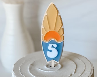Puede incluir: Un pastel blanco con un adorno de pastel en forma de tabla de surf. La tabla de surf es amarilla con un sol naranja y una ola azul. El número 5 está en la ola.