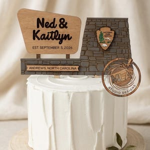 May include: A wooden cake topper featuring a National Park Service sign design with the text Ned & Kaitlyn, EST. SEPTEMBER 5, 2026, and ANDREWS, NORTH CAROLINA. The topper is placed on a white frosted cake with a textured surface and stone wall detail.