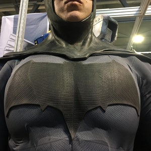 Puede incluir: Primer plano de una persona que lleva un traje de Batman negro. El traje presenta un símbolo de murciélago detallado en el pecho.