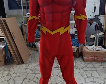 May include: A red and yellow superhero costume with a lightning bolt symbol on the chest. The costume is made of a stretchy fabric and has a muscular design. The costume is on a mannequin and is displayed in a workshop setting.