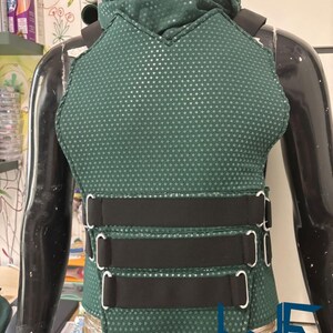 May include: A dark green vest with a textured, dotted pattern and black straps. The vest has a high collar and three black straps with silver buckles across the midsection. The Wayne Factory Costuming Studio logo is visible at the bottom.