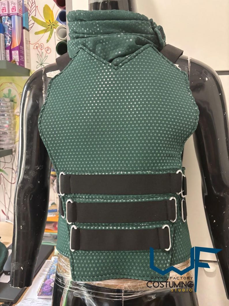 May include: A dark green vest with a textured, dotted pattern and black straps. The vest has a high collar and three black straps with silver buckles across the midsection. The Wayne Factory Costuming Studio logo is visible at the bottom.