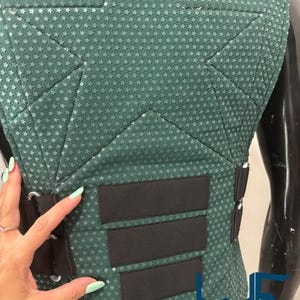 May include: Close-up of a teal-green costume piece with a star pattern. The piece has black straps and accents. The text "WAYNE FACTORY COSTUMING STUDIO" is visible.