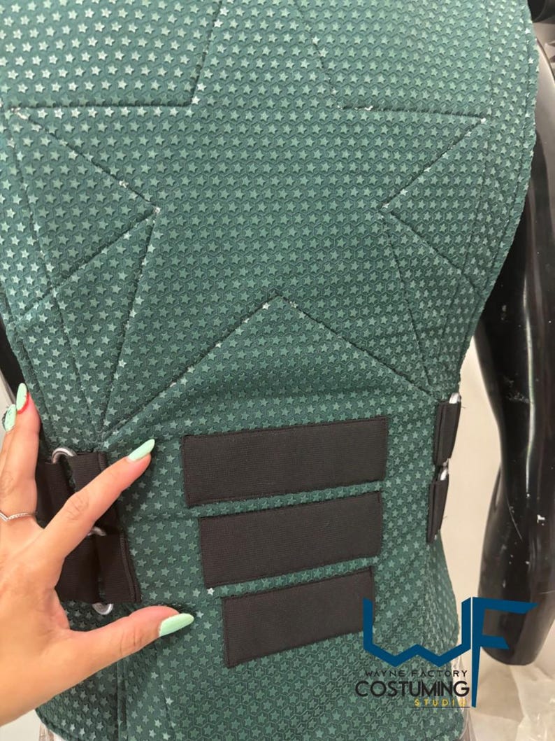 May include: Close-up of a teal-green costume piece with a star pattern. The piece has black straps and accents. The text "WAYNE FACTORY COSTUMING STUDIO" is visible.