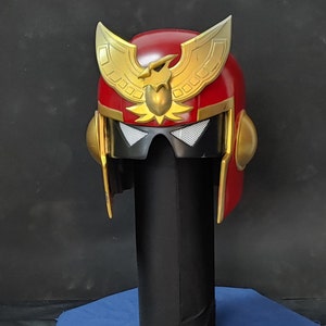 May include: A red and gold helmet with a stylized eagle design on the front. The helmet has a black visor and a gold trim.