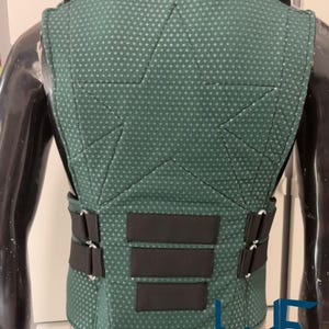May include: A green vest with a textured pattern and black accents. The vest has a high collar and multiple black straps and panels. The back of the vest is visible, showcasing the design and stitching. The vest is displayed on a mannequin.