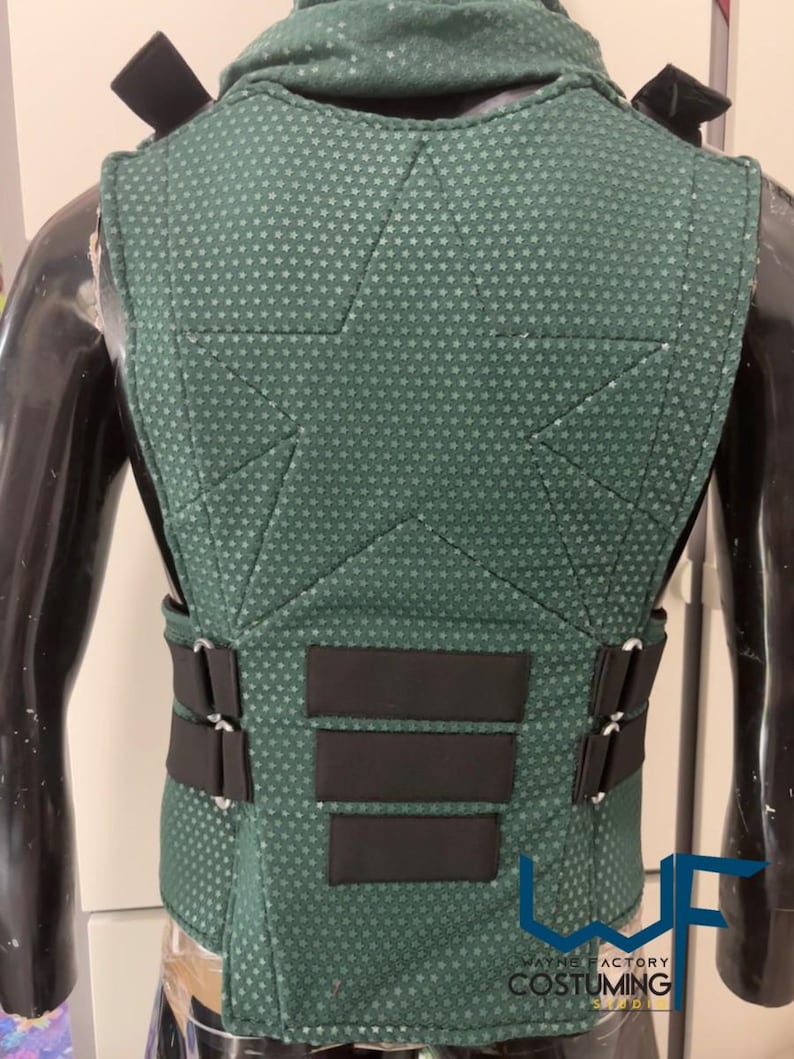 May include: A green vest with a textured pattern and black accents. The vest has a high collar and multiple black straps and panels. The back of the vest is visible, showcasing the design and stitching. The vest is displayed on a mannequin.