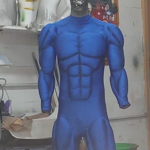 Może przedstawiać: Niebieski, muskularny, jednoczęściowy strój na manekinie. Strój ma srebrną maskę na twarz z czarnym wzorem. Manekin stoi przed białą ścianą z plakatem Marvela w tle.