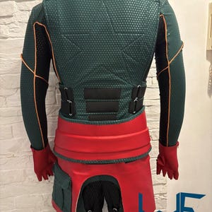 May include: A green and black costume with orange trim, a red belt, and red gloves. The costume has a textured pattern and includes a hood and multiple straps. The costume has the text "WAYNE FACTORY COSTUMING".