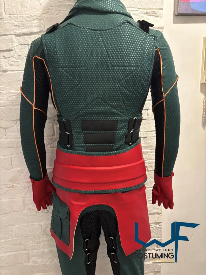 May include: A green and black costume with orange trim, a red belt, and red gloves. The costume has a textured pattern and includes a hood and multiple straps. The costume has the text "WAYNE FACTORY COSTUMING".