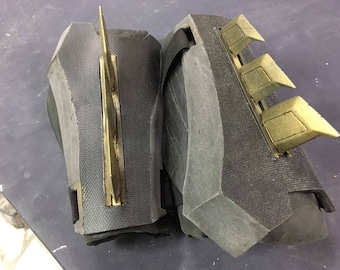 May include: A pair of black foam gauntlets with gold metal accents. The gauntlets have a textured surface and are designed to protect the wearer's hands.