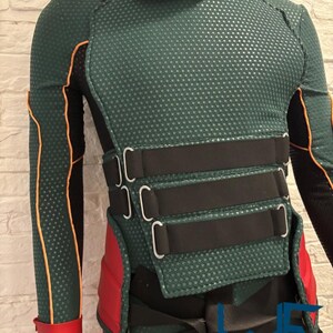 May include: A green and black costume with red accents. The costume features a textured pattern, black straps, and a high collar. The design includes orange trim and a red glove. The costume has the text "COSTUMING STUDIO".