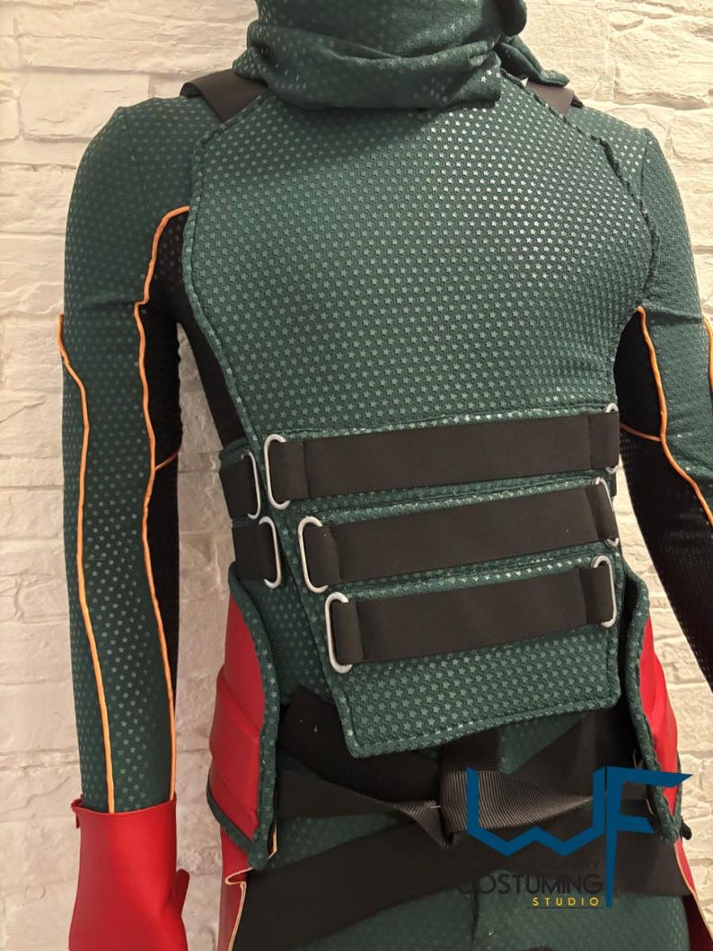 May include: A green and black costume with red accents. The costume features a textured pattern, black straps, and a high collar. The design includes orange trim and a red glove. The costume has the text "COSTUMING STUDIO".