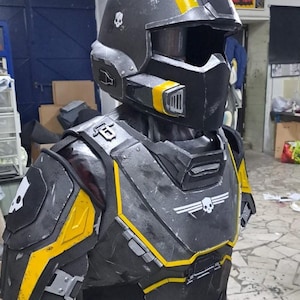 May include: A black and yellow tactical helmet and chest armor set featuring a weathered finish, white skull emblems on the helmet and shoulder, and a white winged skull logo on the chest plate. The gear includes integrated vents and straps.