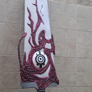 May include: A large, stylized sword prop with a white blade and intricate burgundy-colored organic designs. The hilt is black and wrapped. The blade features a central circular emblem. The text "WAYNE FACTORY COSTUMING STUDIO" is visible.