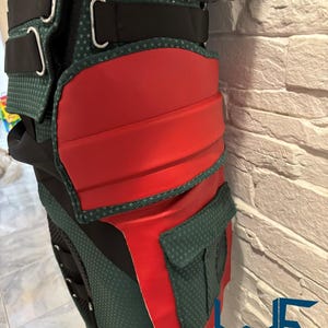 May include: Close-up of a green and red costume piece with black straps and silver buckles. The costume has a textured green fabric with red accents and a small pocket. The logo "WAYNE FACTORY COSTUMING" is visible.