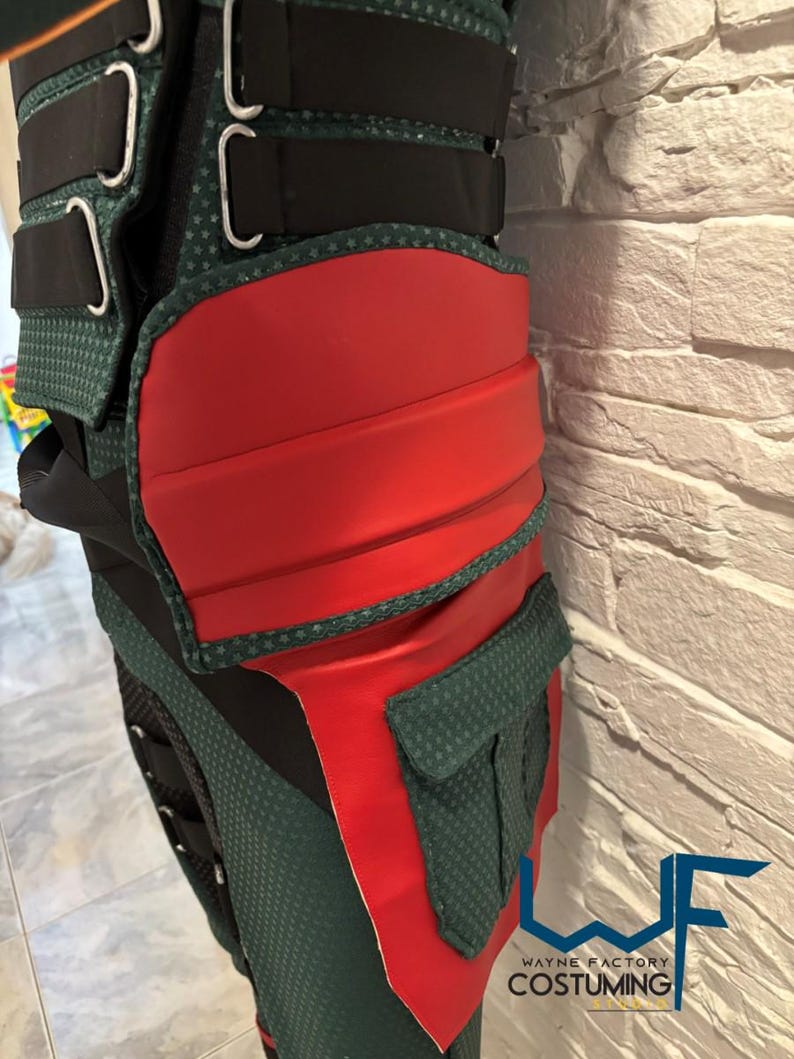 May include: Close-up of a green and red costume piece with black straps and silver buckles. The costume has a textured green fabric with red accents and a small pocket. The logo "WAYNE FACTORY COSTUMING" is visible.
