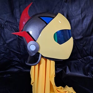 Puede incluir: Un casco amarillo y gris con un diseño rojo y azul. El casco tiene una visera y un cuerno grande y curvo en la parte superior. El casco está sentado sobre un paño amarillo.