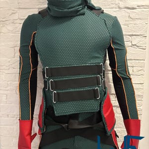 May include: A green and black costume with orange trim, red gloves, and a high collar. The costume features a textured pattern and black straps across the chest and waist. The costume is displayed on a mannequin.