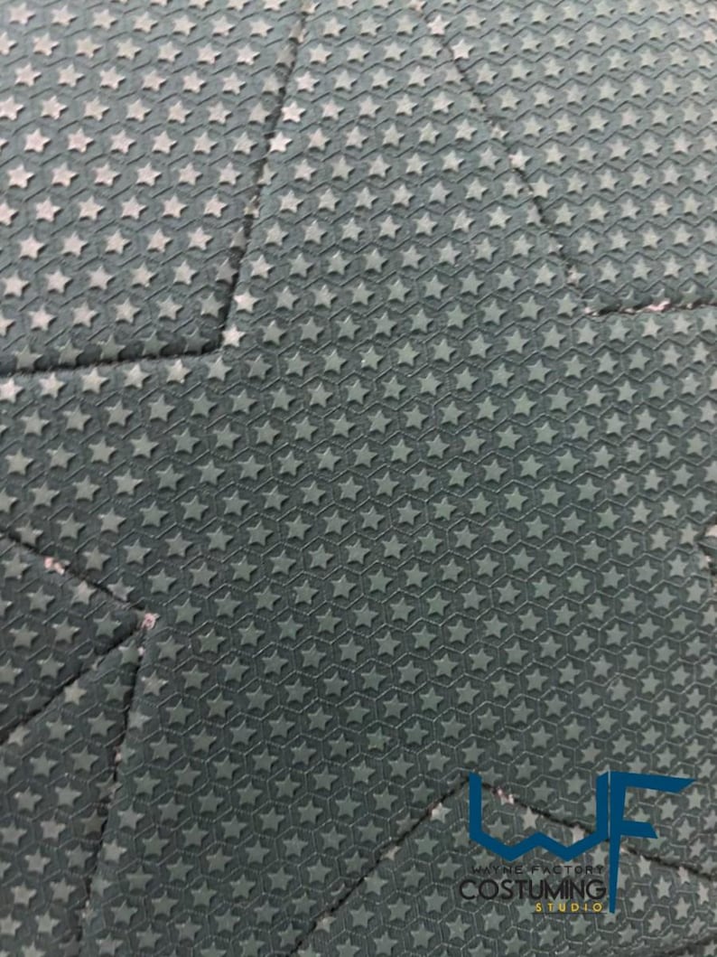 May include: Close-up of a dark teal fabric with a repeating pattern of small, raised stars. The fabric is divided into geometric sections by seams. The lower right corner features a logo with the text "WAYNE FACTORY COSTUMING STUDIO."