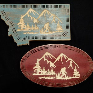 May include: Two wooden cribbage boards with a mountain and Bigfoot design. One board is shaped like the state of Montana and is painted blue. The other board is oval and is painted red.