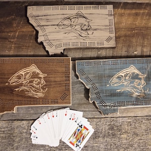 May include: Three wooden cribbage boards shaped like the state of Montana, each with a carved fish design. One board is brown, one is blue, and one is gray. A deck of playing cards is fanned out in front of the boards.