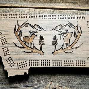 May include: A light-colored wooden cribbage board shaped like the state of Montana. The surface features laser-engraved imagery of two large antlers framing a deer, an elk, pine trees, and mountain peaks, with rows of drilled peg holes.